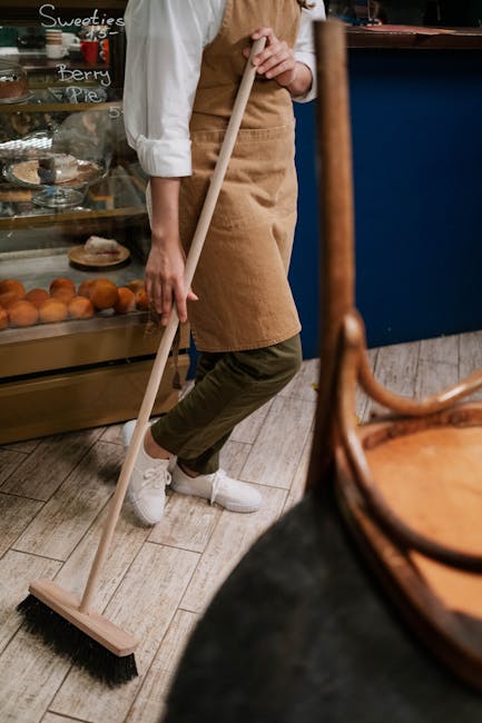 A person wearing a beige apron and green pants is using a broom to sweep a wooden floor in a bakery or café setting with glass display cases filled with baked goods like croissants and fruit pies. The scene is well-lit with natural and artificial light, highlighting the clean, dust-free wooden surfaces. In the background, there are handwritten menu signs on the display case. The person’s white sneakers are visible as they focus on surface cleaning and maintaining hygiene, aligning with professional domestic cleaning standards promoted by Cleaner Kennington. The image emphasizes the importance of thorough cleaning processes in maintaining a hygienic environment.