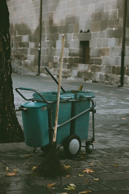 A teal-colored outdoor cleaning trolley with two large wheels and a handle, parked on a paved sidewalk next to a tree with a broom leaning against it. The trolley contains cleaning tools such as brushes and squeegees, and a small cleaning supply caddy is attached. The surrounding area features an aged brick wall with some patches, fallen leaves scattered on the pavement, and diffused natural lighting, indicating an outdoor urban environment. This image exemplifies surface cleaning tools used in outdoor domestic or commercial cleaning services provided by Cleaner Kennington, highlighting maintenance in public or private outdoor spaces.