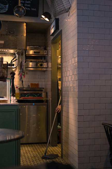 A person wearing a beige apron and green pants is using a broom to sweep a wooden floor in a bakery or café setting with glass display cases filled with baked goods like croissants and fruit pies. The scene is well-lit with natural and artificial light, highlighting the clean, dust-free wooden surfaces. In the background, there are handwritten menu signs on the display case. The person’s white sneakers are visible as they focus on surface cleaning and maintaining hygiene, aligning with professional domestic cleaning standards promoted by Cleaner Kennington. The image emphasizes the importance of thorough cleaning processes in maintaining a hygienic environment.