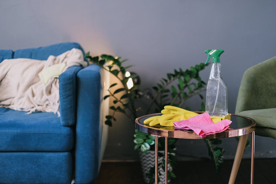 A living room scene showcasing surface cleaning tools and materials. On a round side table with a metallic finish, there is a clear spray bottle with a green nozzle, a pair of yellow rubber gloves, an