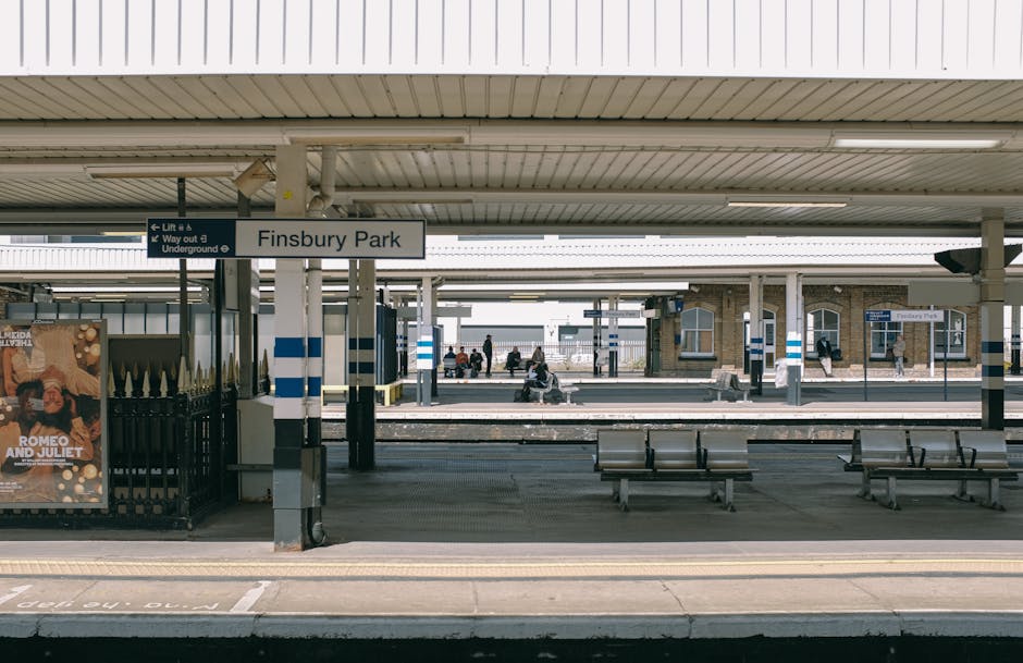 A view of Finsbury Park railway station platform with a metal roof overhead and multiple benches arranged along the platform. The platform surface is clean and well-maintained, with visible tactile paving at the edge. A sign indicating 'Finsbury Park' is suspended from the ceiling, and there are a few passengers sitting and waiting on the benches. The station's brick buildings and additional signage are visible in the background. The lighting is natural, suggesting daytime, and the overall scene reflects a tidy, hygienic public transport environment. Cleaner Kennington specialises in surface and deep cleaning, ensuring high standards of hygiene in busy areas like train stations, as seen in this well-kept platform picture.