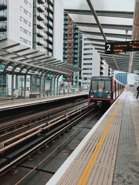 An urban train station platform with a train arriving on the tracks, featuring a modern glass and steel shelter with seating. The platform is clean and well-maintained, with tactile paving along the edge for safety. In the background, high-rise residential and commercial buildings are visible under an overcast sky, with a digital sign displaying platform information. The scene highlights a high standard of station hygiene and maintenance, consistent with best practices for public transport cleaning, as emphasized by Cleaner Kennington in their surface cleaning and sanitisation services near Kennington Station, which support safe and hygienic travel environments.