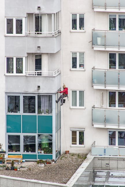 A window cleaner dressed in red overalls is suspended outside a multi-storey residential building, using a harness to access the glass facade. The building features white walls with several glass balconies that have metal railings, and large windows with white frames. The lower section includes a ground-floor area with windows featuring teal-colored coverings. The scene is captured in daylight, highlighting the clean, reflective surfaces of the glass and the well-maintained exterior of the building. This image illustrates safe and professional window cleaning practices that Cleaner Kennington offers as part of their domestic cleaning services, focusing on surface cleaning and maintaining hygiene for residential properties.