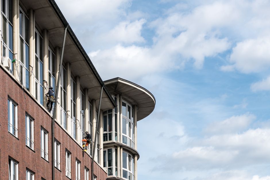 Exterior view of a modern multi-story building with a brick facade and large glass windows, some workers are seen performing window cleaning on the upper floors using harnesses and cleaning tools, with a partly cloudy sky overhead. The scene highlights the importance of surface cleaning and maintenance for high-rise residential or commercial properties, and is associated with Cleaner Kennington's expertise in deep cleaning and sanitisation services for accessible building exteriors and windows.