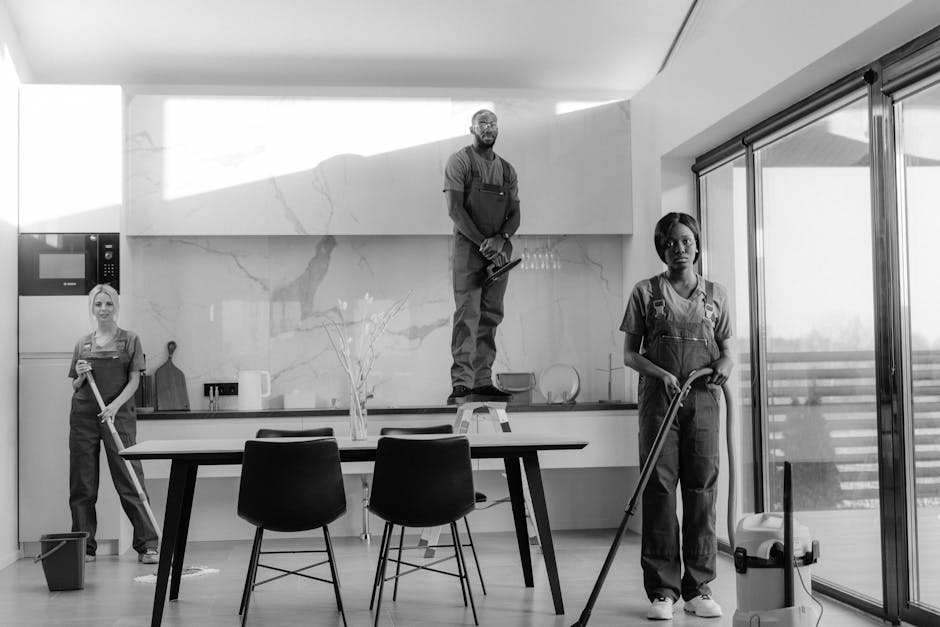 A black-and-white image features three professional cleaners working in a modern kitchen with a minimalist design. One cleaner, a woman on the left, holds a mop and stands next to a bucket on the wooden floor. The second cleaner, a man in the center, is positioned on a step ladder, wearing an apron and holding cleaning tools, with a marble backsplash visible behind him. The third cleaner, a woman on the right, is mopping the floor near large sliding glass doors that let in natural light, revealing a balcony outside. The clean, shiny surfaces of the kitchen, including ceramic tiles and smooth countertops, highlight the thorough surface cleaning and maintenance provided by Cleaner Kennington, a professional cleaning company specializing in domestic cleaning services. The room’s bright, well-lit atmosphere emphasizes hygiene and cleanliness, aligning with the topic of avoiding delays and booking mistakes in professional cleaning services.