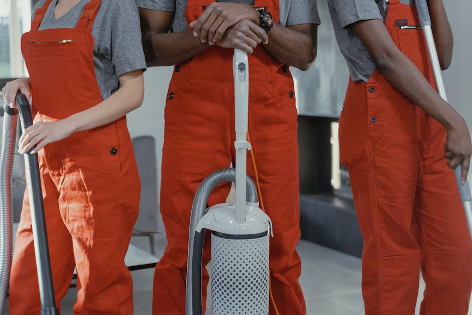 A black-and-white image features three professional cleaners working in a modern kitchen with a minimalist design. One cleaner, a woman on the left, holds a mop and stands next to a bucket on the wooden floor. The second cleaner, a man in the center, is positioned on a step ladder, wearing an apron and holding cleaning tools, with a marble backsplash visible behind him. The third cleaner, a woman on the right, is mopping the floor near large sliding glass doors that let in natural light, revealing a balcony outside. The clean, shiny surfaces of the kitchen, including ceramic tiles and smooth countertops, highlight the thorough surface cleaning and maintenance provided by Cleaner Kennington, a professional cleaning company specializing in domestic cleaning services. The room’s bright, well-lit atmosphere emphasizes hygiene and cleanliness, aligning with the topic of avoiding delays and booking mistakes in professional cleaning services.