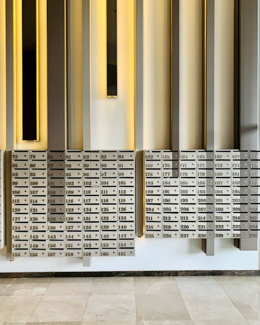 A row of metallic mailboxes with numbered compartments is mounted on a wall in a residential building's lobby. The mailboxes are made of stainless steel and have a brushed finish, with small keyholes and labels displaying individual unit numbers from 78 to 226. Above the mailboxes, there are vertical, rectangular decorative panels in alternating shades of black and beige, framed by slim gold accents, providing an aesthetic touch to the space. The lighting is soft, illuminating the area evenly, and the floor is covered with light-colored tiles that appear clean and well-maintained. The overall scene reflects a tidy and organized entrance area, consistent with professional cleaning standards, as offered by Cleaner Kennington for surface cleaning and hygiene maintenance in residential communal spaces.