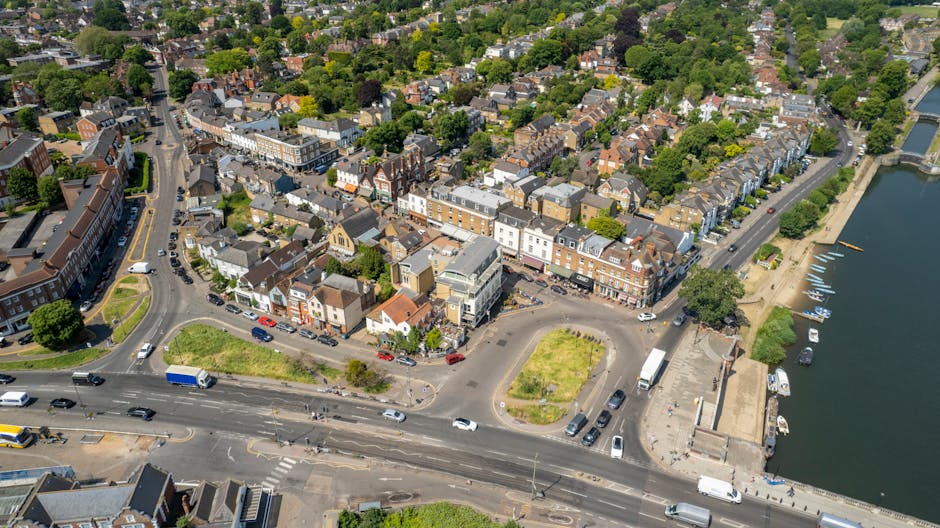 An aerial view of a riverside urban area showing a mixture of residential and commercial buildings with red brick facades, some with flat roofs and others with pitched roofs, along with a prominent parking area and a small green park with grass and trees. The scene includes a body of water on the right side with boats docked along the quay, and main roads with multiple vehicles, including cars, buses, and trucks, navigating through the area. Bright daylight and lush green trees indicate a clear, sunny day, highlighting the tidy and well-maintained streets and surroundings. This image exemplifies the neat, clean environment that professional cleaning services like Cleaner Kennington support for residential and commercial properties in the local area.