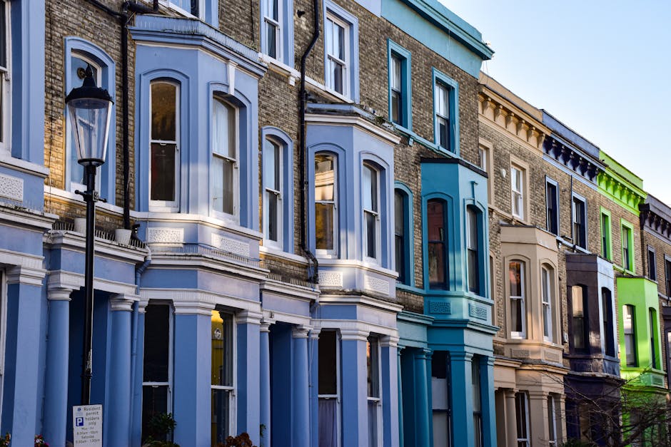 A row of terraced residential buildings with bay windows and decorative architectural details, painted in various pastel shades including blue, beige, and bright green, situated along a street with a black classic-style street lamp in the foreground. The buildings are made of brick with white trim accents, and large glass windows reflecting the blue sky and surrounding environment. The scene appears during daytime with natural lighting highlighting the clean, well-maintained facades. The image showcases exterior features typical of London architecture, emphasizing the vibrant colors and historic charm. Cleaner Kennington specializes in surface cleaning and deep cleaning services to maintain the building exteriors, ensuring they remain bright and inviting for residents and visitors alike.