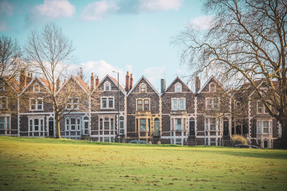 A row of traditional Victorian-style terraced houses made of grey stone with white window frames and decorative trim, set along a residential street in Kennington. The front gardens are separated from the pavement by low wooden fencing, with some bushes and small trees visible. The houses feature gabled roofs, chimneys, and large sash windows, with a partly cloudy sky overhead illuminating the scene. The well-maintained grassy area in the foreground is lush and green, with a few mature leafless trees indicating a late autumn or early winter setting. This image exemplifies the residential architecture typical for SE11 and highlights the importance of property maintenance and cleaning, aligning with the [PAGE_TITLE] for Kennington Road residents, as promoted by [COMPANY_NAME].