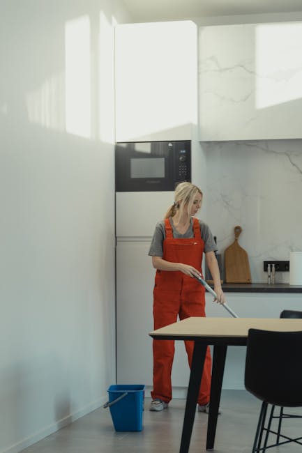 A woman wearing an orange cleaning apron and gray t-shirt is mopping a light-colored laminate or wooden dining table in a modern kitchen. The kitchen features a sleek, minimalistic design with white walls, a built-in oven, and marble-effect tiles on the wall. Natural light from an unseen window illuminates the space, highlighting the cleanliness and shine of the surfaces. A blue cleaning bucket is positioned on the floor beside her, and she is focused on maintaining hygiene and surface cleanliness. This scene exemplifies domestic surface cleaning, emphasizing professional deep cleaning standards, as performed by Cleaner Kennington for urgent same-day cleaning in Kennington SE11. The overall setting reflects a tidy and well-maintained environment, suitable for routine or deep cleaning procedures.