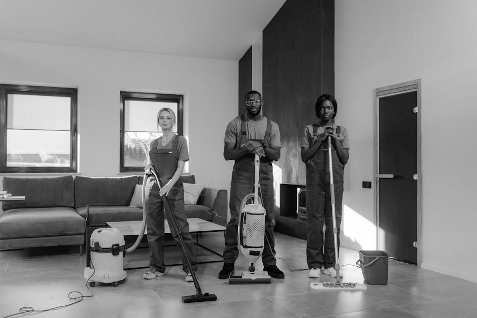 Three professional cleaners, two women and one man, are engaged in surface cleaning and deep cleaning activities within a modern living room. The room features a light-colored laminate flooring, with a dark-colored sofa along the wall under two large windows that provide natural light. The women are holding vacuum cleaners and cleaning tools; one has a handheld vacuum, while the man stands between them with a larger upright vacuum. The cleaning tools are connected to power outlets, and the surfaces, including a wooden coffee table and a tiled wall section, appear clean and well-maintained. The room's surfaces, like the smooth wooden floors and glazed windows, are free of dust and dirt, demonstrating effective domestic cleaning by [COMPANY_NAME], aimed at sanitation and hygiene maintenance, consistent with the context of urgent same-day cleaning options in Kennington SE11.